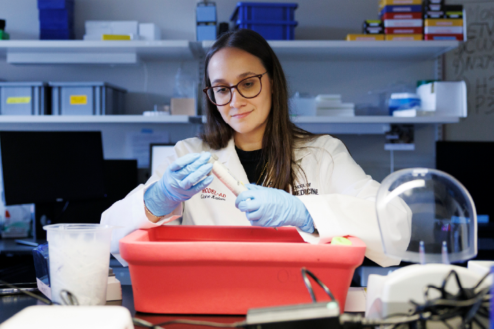 A woman in a lab coat works with samples while wearing blue latex gloves over a large red container in a laboratory