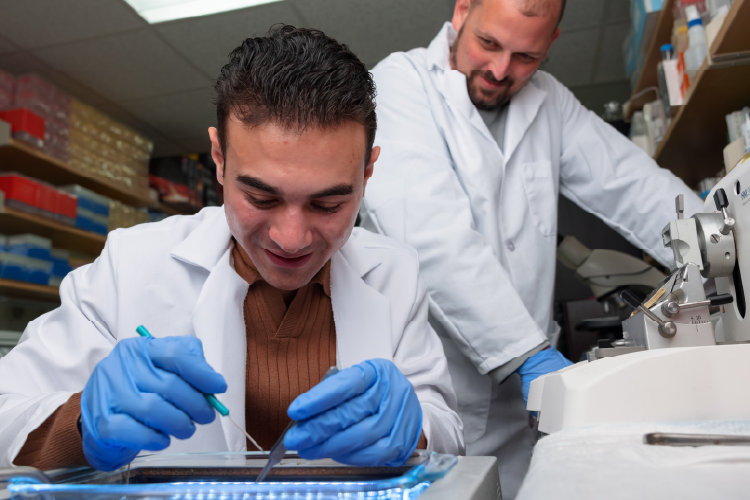 A student in a white lab coat and blue exam gloves. He is watched by an mentor, also dressed in a white jacket, in a laboratory setting.