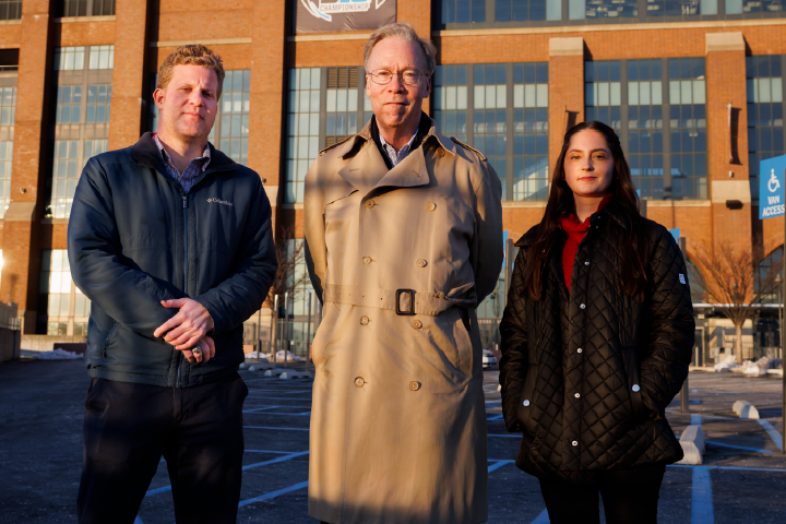 Two students and their professor dressed in winter coats stand outside of Lucas Oil Stadium in Indianapolis, Indiana during the day
