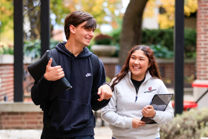 Two students holding a tablet and laptop case walk together through a brick surrounded courtyard.