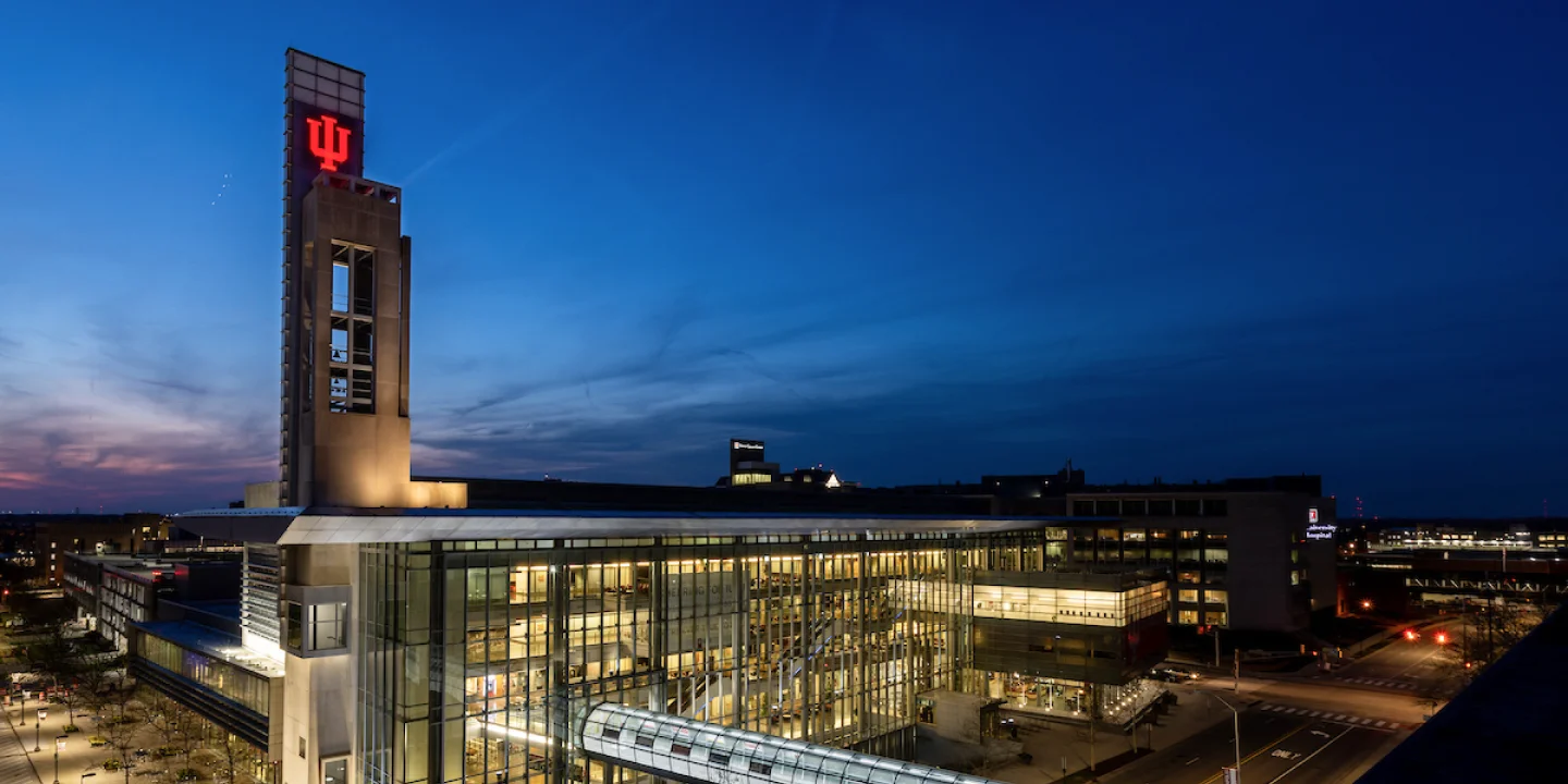 The Campus Center is seen lit up from inside at night. A red trident glows from the bell tower and the night sky is going from a pink to a dark blue as the sun sets.