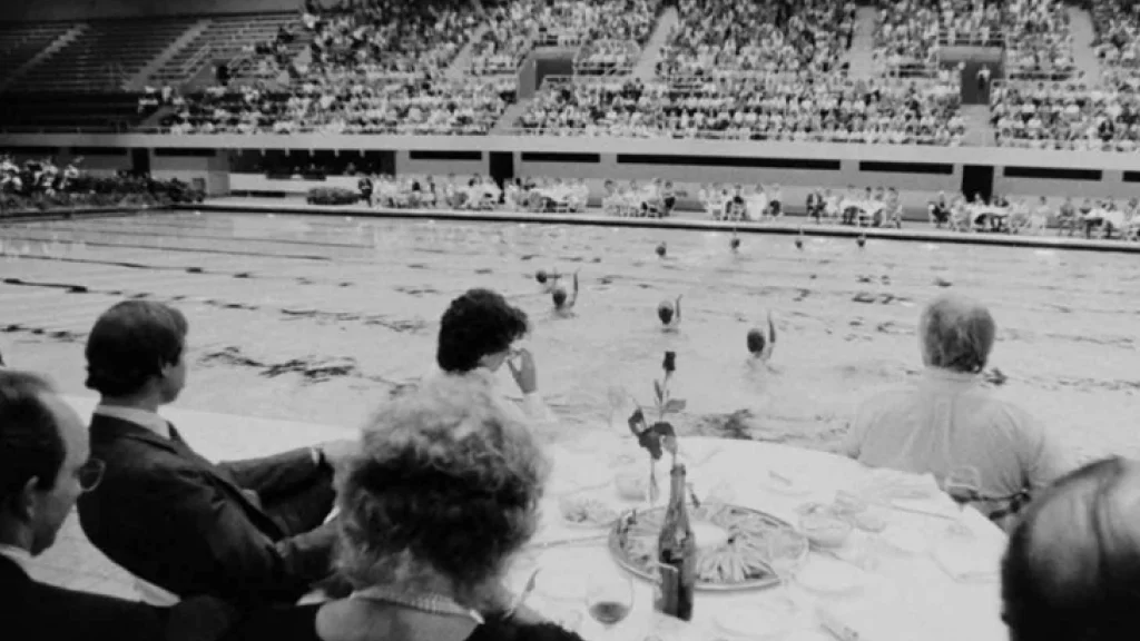 Spectators around a table watch as student swim in the 1983 Classic Splash in the Natatorium.