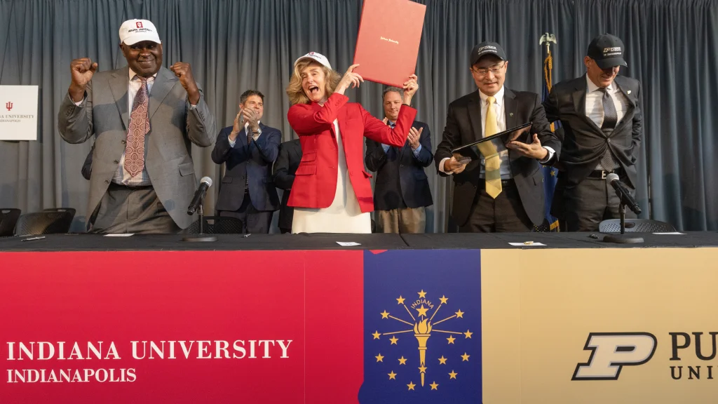 President Pamela Whitten holds up a leather folio as board of trustees members and Purdue leadership clap following the joint agreement signing ceremony that officially creates separate and independent Indianapolis campuses.