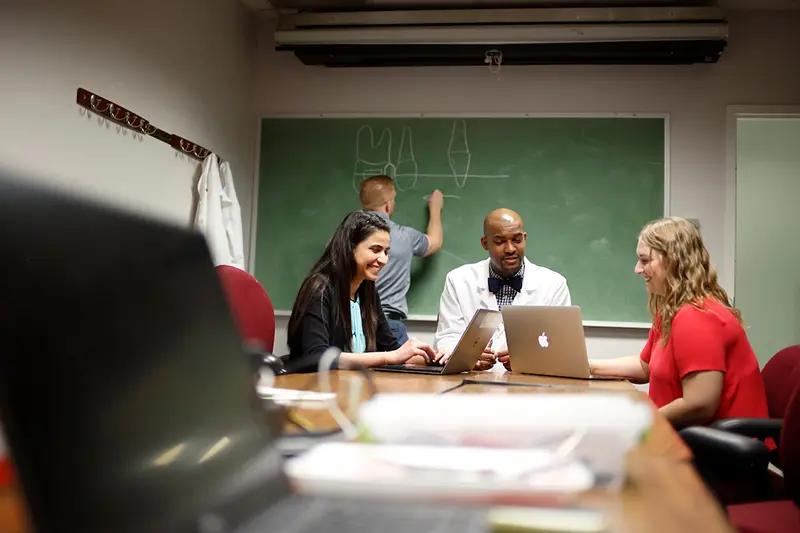 Students and professor sitting at table with laptops while someone writes on the chalkboard behind them