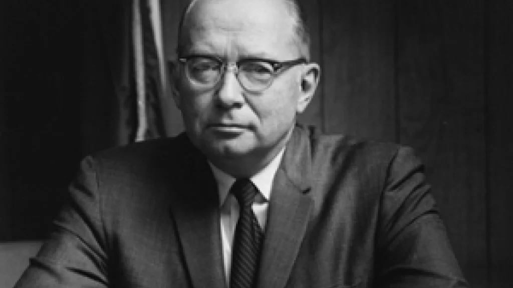 Chancellor Maynard K. Hine sits swearing a suit, hand interlocked, in front a wood paneled wall.