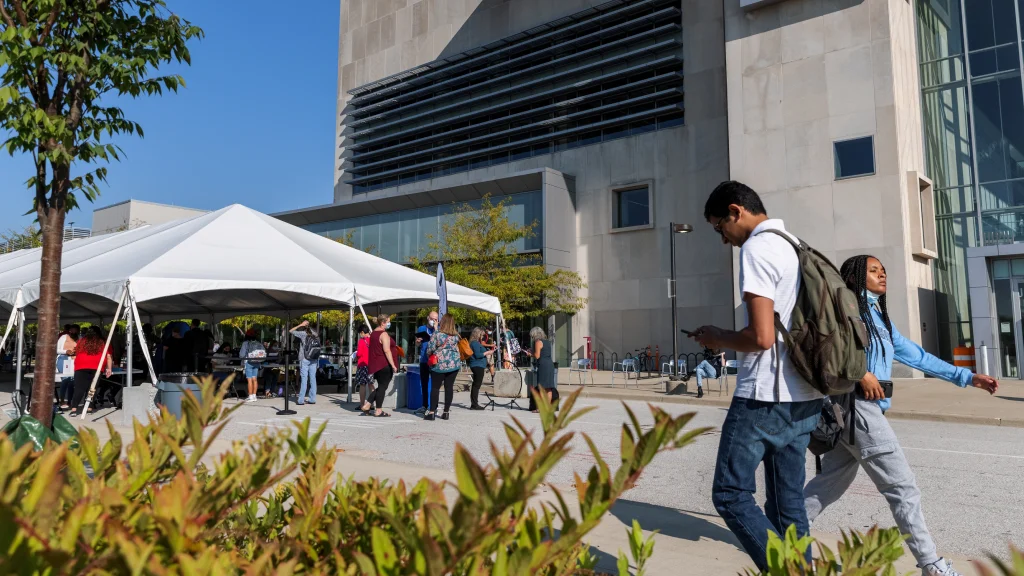 Two students pass by walking outside the campus center. A white event tent is set up in the street to the left for an event and landscaping is seen in the foreground.