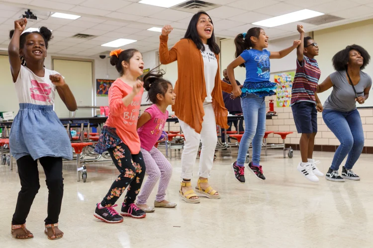 Two teachers and five children gather in a line, some in mid-jump, during an elementary classroom activity.