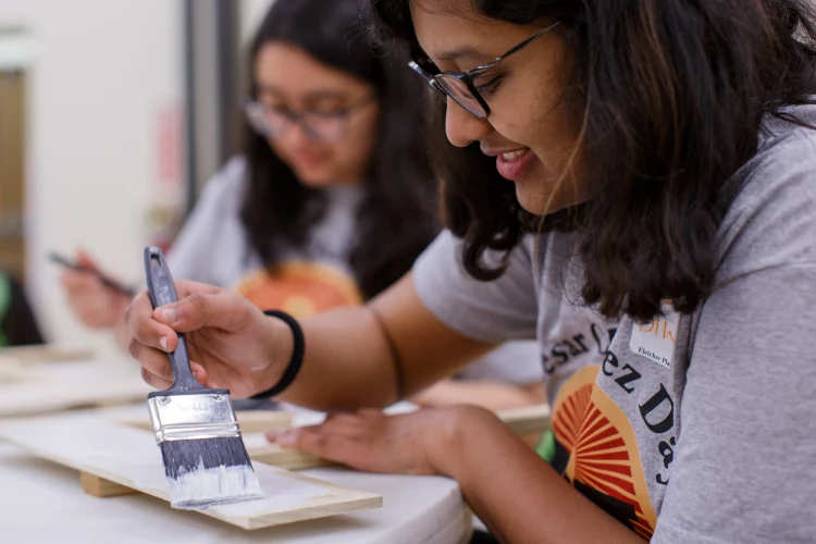 Two students paint pieces of wood resting on a table with white paint on handheld brushes.