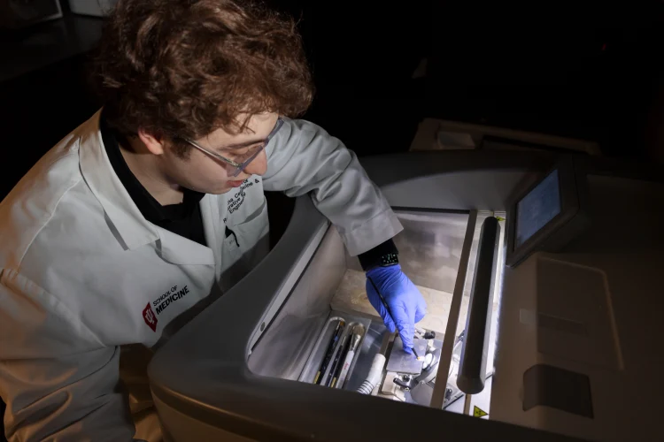A student in a lab coat and medical gloves reaches inside a piece of machinery lit up from inside to show parts and technology.