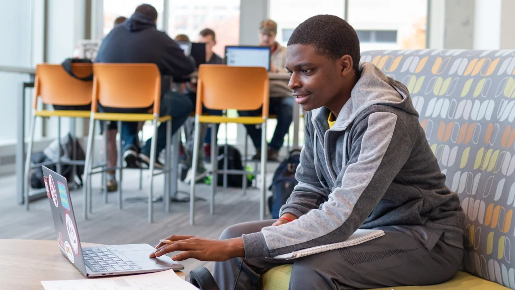 A student sits on an upholstered couch using a laptop in front of him. Other students sit at a high table in a common area behind him.