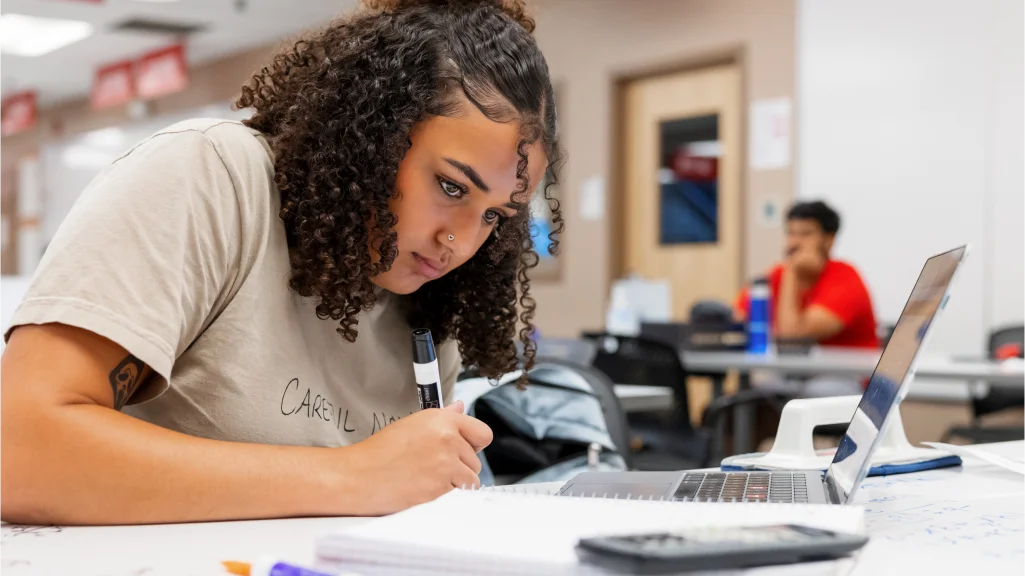 A student sits at a table with a laptop, notebook, calculator and dry erase markers.