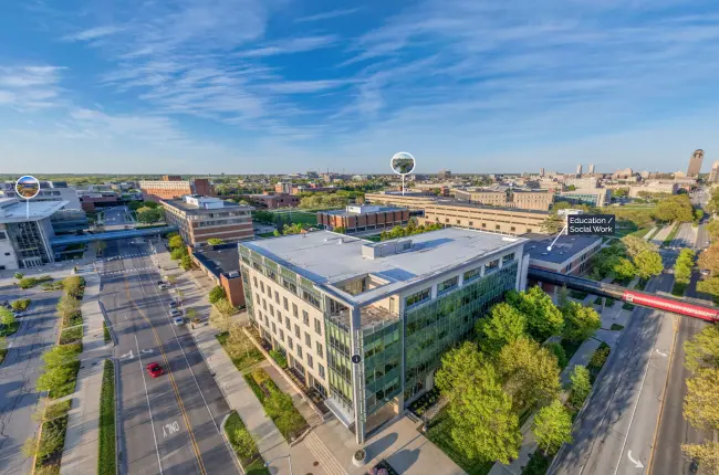 Aerial view of modern university buildings with large glass windows, situated on tree-lined streets with adjacent academic buildings and green spaces and the city skyline visible in the distance.