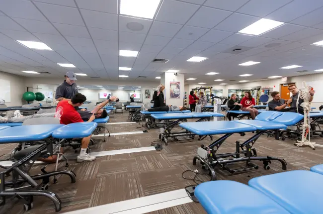 Students practicing physical therapy techniques on treatment tables in a large clinical classroom.