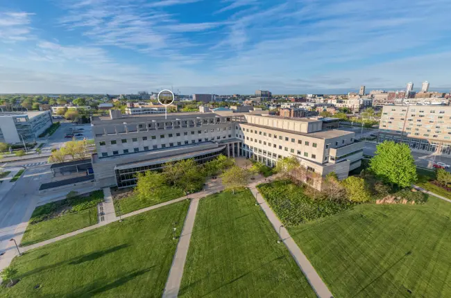 Aerial view of a university campus building with modern architecture with large windows surrounded by green lawns and walkways, with a backdrop of city buildings.