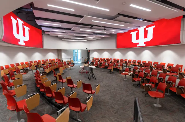 Modern classroom with rows of bright red chairs with attached desks arranged in a circle around a center lectern. IU logos are displayed on large screens on the walls above the chairs.