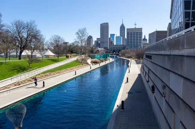 Scenic canal with walkways on both sides, bordered by green lawns and trees, with the Indianapolis city skyline in the background.