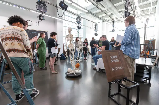 Art students gathered around skeleton models in a well-lit studio with large windows and high ceilings.