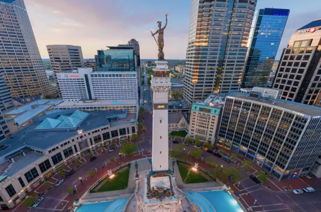 Aerial view of downtown Indianapolis at dusk, showcasing the iconic Soldiers and Sailors Monument with its detailed sculpture at the top, surrounded by illuminated skyscrapers.