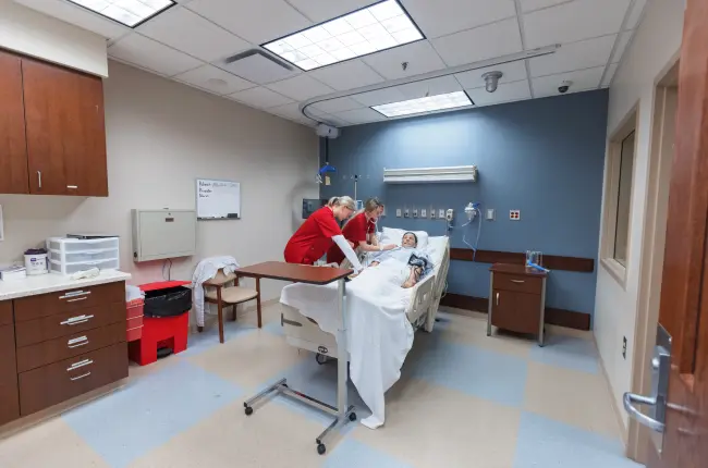 Nursing students in red scrubs practicing on a patient simulator in a clinical training room.