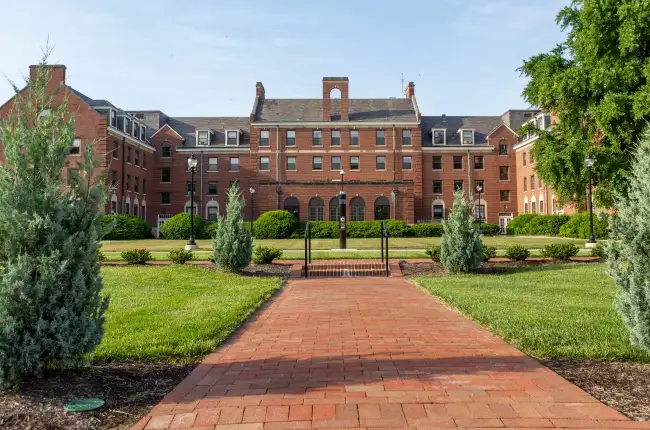 Four story red-brick university building with trees and landscaping in the foreground, with a paved brick sidewalk leading toward the building.