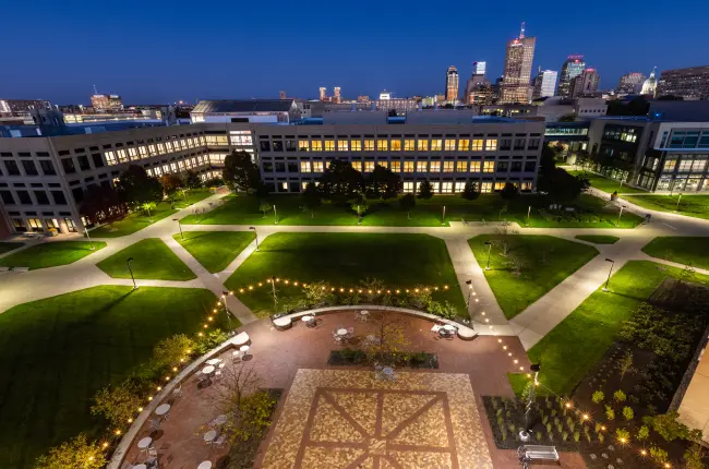 Illuminated university courtyard with walkways and buildings, set against a backdrop of a city skyline.