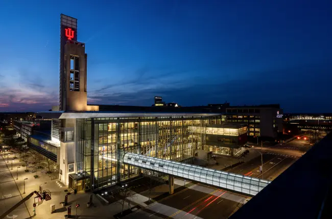 Modern building with a skywalk and illuminated IU logo on a tower.