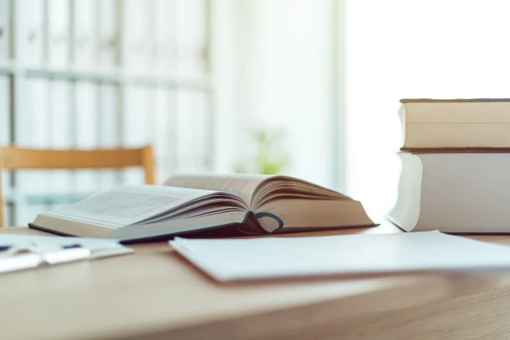 An open book and two stacked books sit on a wooden desk in a bright, airy room, with shelves seen in the background.