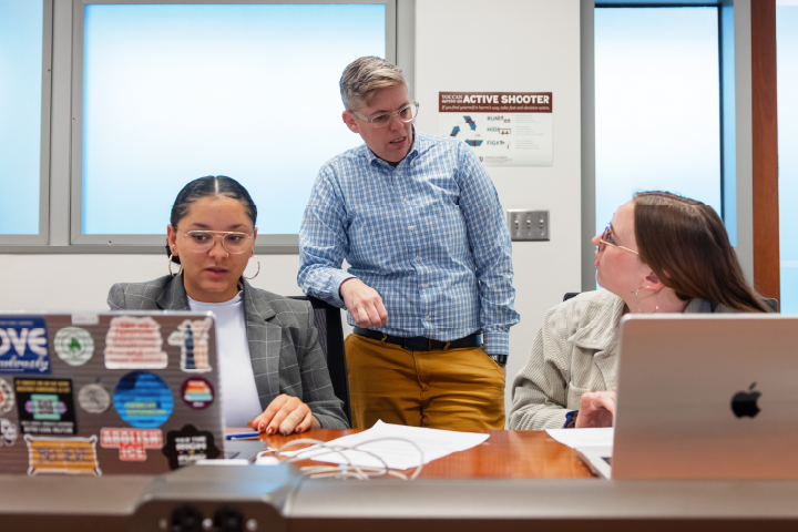 Two students sitting at laptops along with mentor standing behind them discussing.