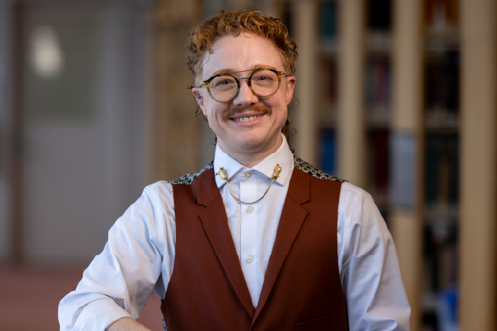 Person smiling at the camera and wearing a brick colored vest and glasses.