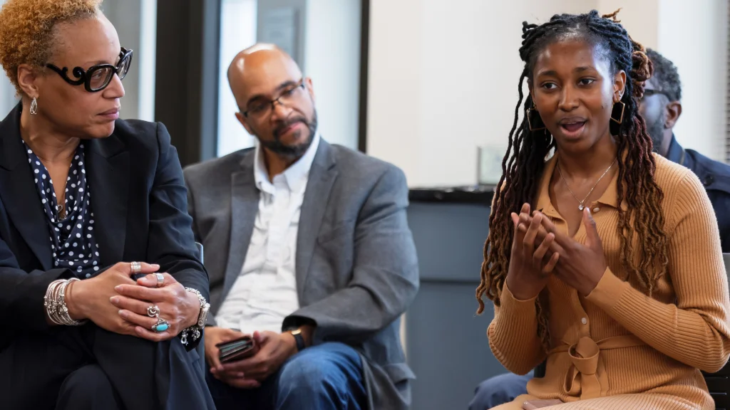 A woman sits among three other people listening attentively, gesturing as she speaks, her hands held in front of her heart.