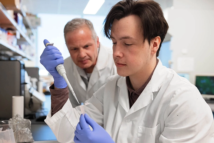 A student in a white lab coat and blue exam gloves uses a pipet to in a laboratory. He is watched by an older mentor, also dressed in a white jacket, in a laboratory setting.