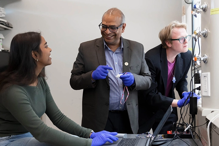 A woman in a green shirt works at a laptop connected to an electronic device while being instructed by a man in a plaid red shirt and black suit jacket.