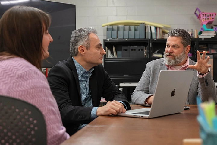 Multiple people in suit jackets and collared shirts sit at a conference table discussing a serious issue.