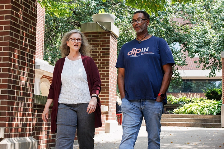 A woman in a white shirt and red cardigan walks outside in front of a brick wall and greenery with a man in a blue t-shirt.