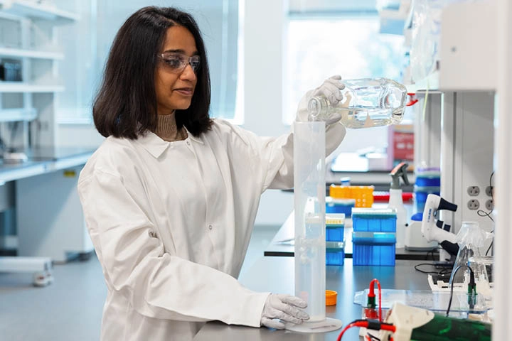 A woman wearing a white lab coat in brightly lit laboratory pours a clear liquid from a glass container into a tall graduated cylinder.