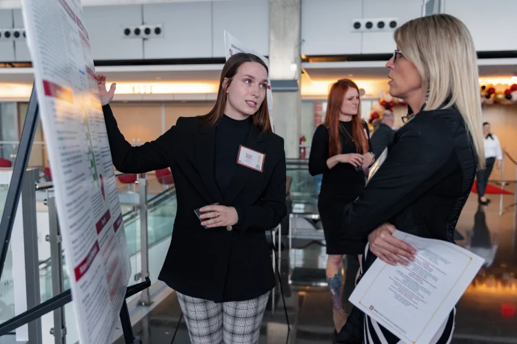 A student with nametag on gestures to a poster displayed on a stand while speaking to a woman at a professional event.