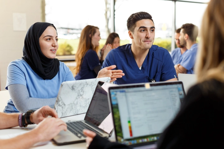Two students face a group of additional students, all with laptops open in front of them, gathered around a table talking.