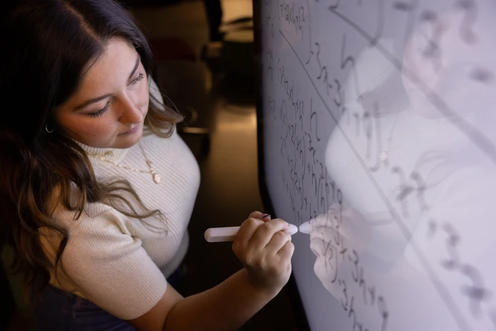 A woman leans in toward a wall display, writing numbers on the screen with an electronic pen.