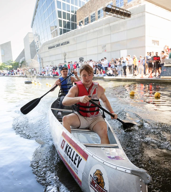 Two students paddling a canoe during the Regatta, wearing life vests, with the front student looking determined. The canoe has a “Kelley School of Business” logo and other sponsor stickers. Spectators line the canal, and the Indiana State Museum is visible in the background. The event appears to be well-attended, with people watching from the side of the canal.