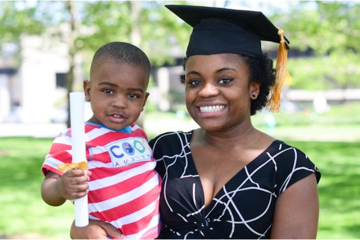 A parent poses at graduation with their young child, who is holding a rolled diploma.