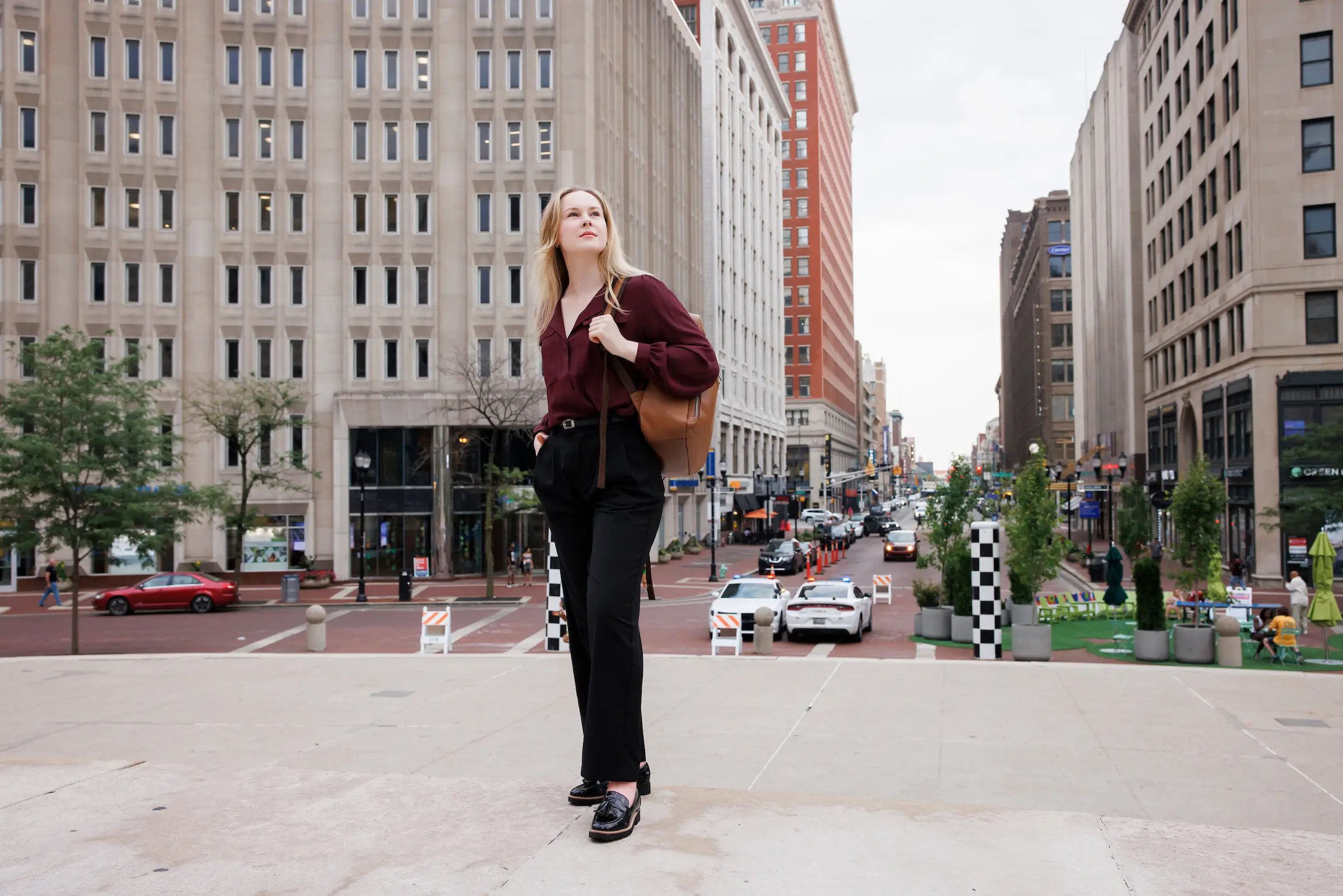 A woman wearing a red blouse and black pants stands in a plaza in downtown Indianapolis. She gazes upwards with tall buildings behind her.