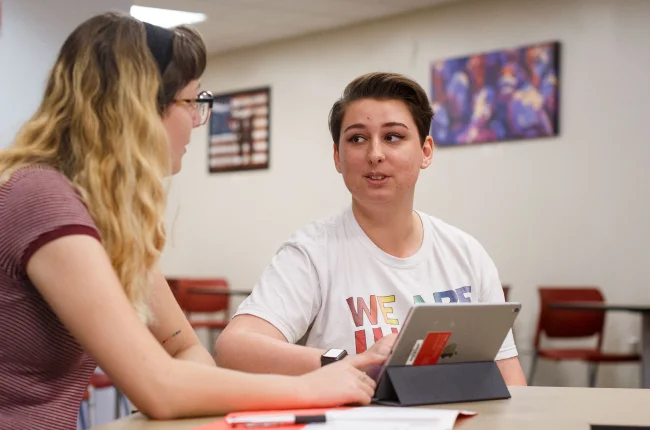 Two students sit at a table with a table and notebook in front of them.