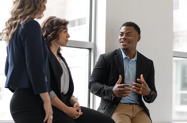 Three people wearing blazers talking in front of large windows