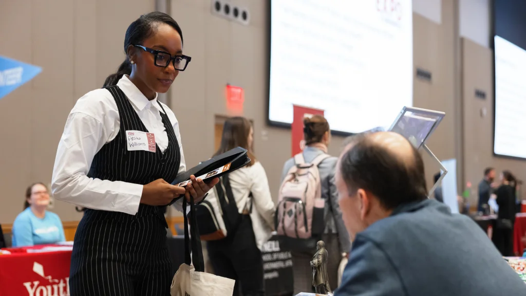 A professionally dressed woman with chunky glasses and laid hair engaging with booth staff at the job fair.