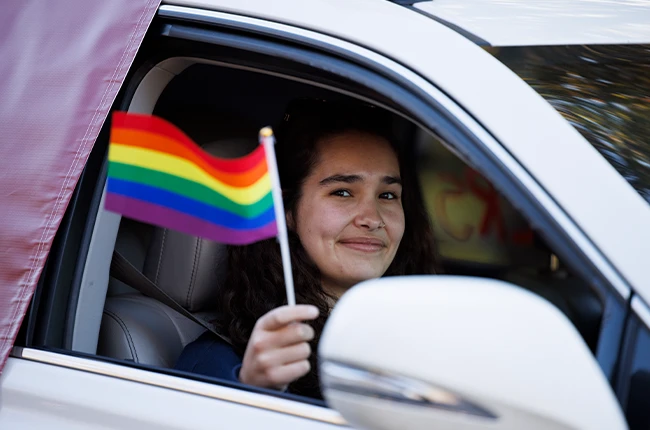 Smiling person in the passenger seat of a car holding a small rainbow flag outside the window