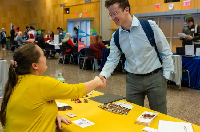 A student leans over a table to shake the hand of a woman seated.