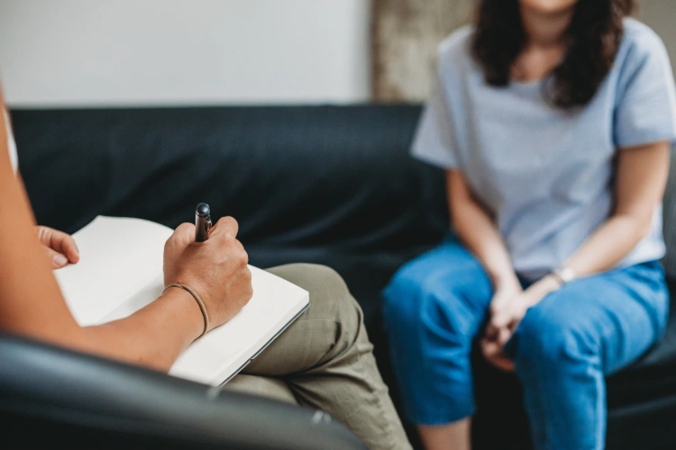 A hand resting on a notepad on a lap takes notes while facing a person sitting across from them on a couch during an appointment.
